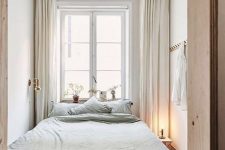 a small and narrow neutral bedroom with a bed with light green bedding, a nightstand with a lamp and neutral drapes for peaceful sleeping