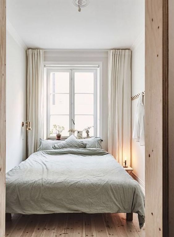 a small and narrow neutral bedroom with a bed with light green bedding, a nightstand with a lamp and neutral drapes for peaceful sleeping