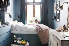a small bedroom in blues, with a platform bed, a dresser, a shabby chic vanity, a pink pouf and some lamps and art