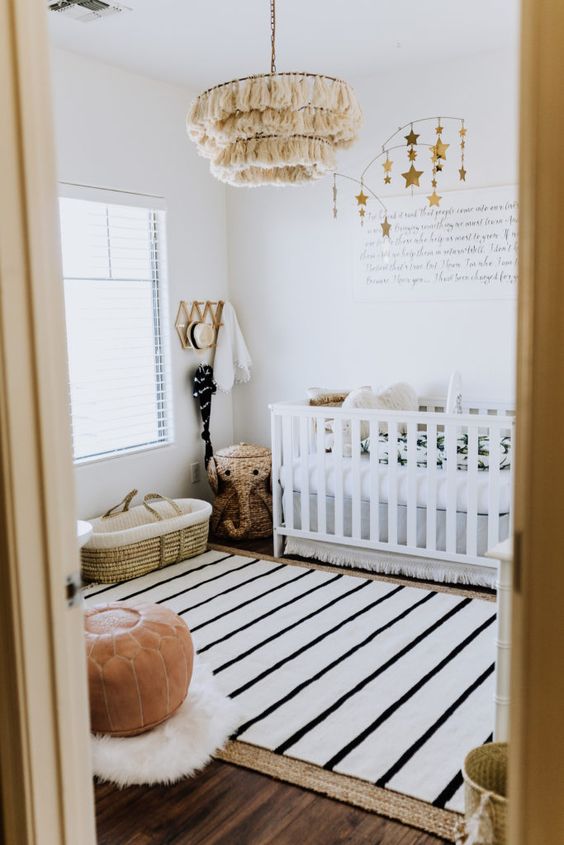 a small boho nursery with layered rugs, a white crib with printed bedding, a leather pouf, tassel chandelier and lovely decor
