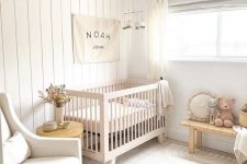 a small neutral nursery with a stained crib, a woven bench, a neutral chair, an ottoman, layered rugs and pretty decor