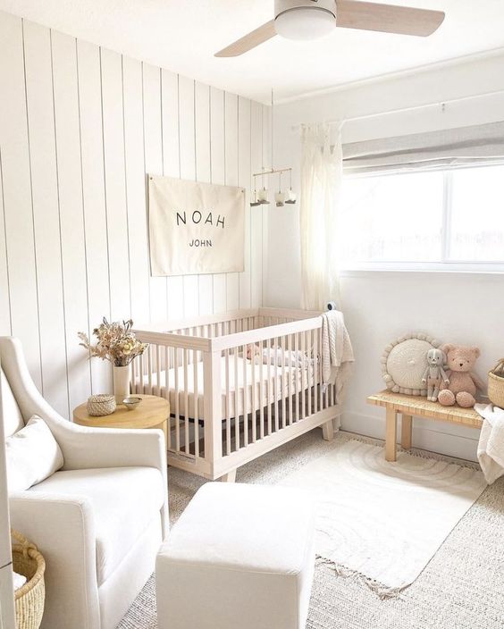 a small neutral nursery with a stained crib, a woven bench, a neutral chair, an ottoman, layered rugs and pretty decor
