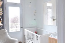 a small neutral nursery with a white crib, a changing table, a neutral chair, some bookshelves and neutral decor