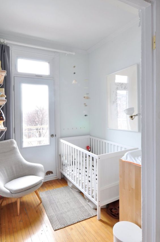 a small neutral nursery with a white crib, a changing table, a neutral chair, some bookshelves and neutral decor