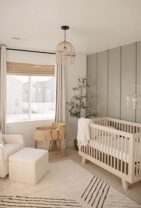 a small neutral nursery with an olive green accent wall, stained crib, a neutral chair, a basset, a printed rug and shades