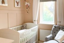 a small pastel nursery with blush walls, a white crib, a grey chair, a gallery wall, some decor and layered drapes