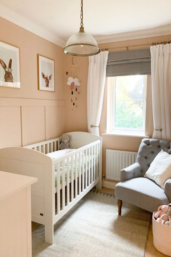 a small pastel nursery with blush walls, a white crib, a grey chair, a gallery wall, some decor and layered drapes