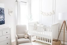 a small white nursery with paneled walls, a white crib, a white dresser, layered rugs, a grey chair with an ottoman and some decor