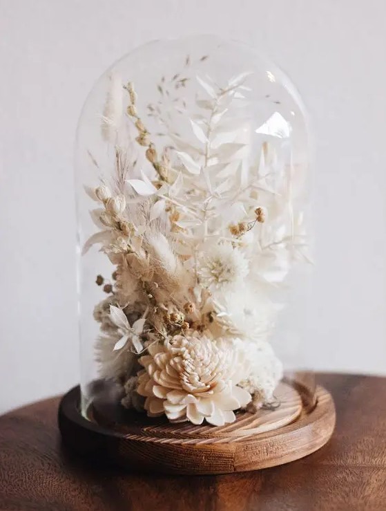 a stylish neutral dried flower arrangement of neutral dried blooms and leaves placed into a cloche with a wooden base