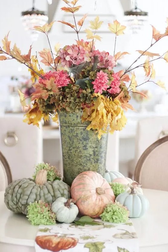 a stylish rustic fall decoration of a bucket with pink and green faux blooms, branches with leaves and heirloom pumpkins