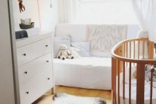 a tiny and cozy mid-century modern nursery with a white dresser, a white sofa, a stained crib, shelves, rugs and neutral curtains