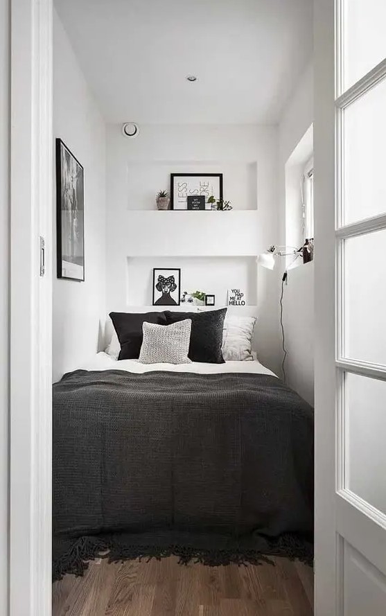 a tiny bedroom in white, with only a bed squuzed in, black and white bedding, niche shelves with decor, an artwork and a sconce