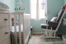 a tiny modern nursery with light blue walls, a white crib and a dresser, a grey chair, a gallery wall and printed textiles