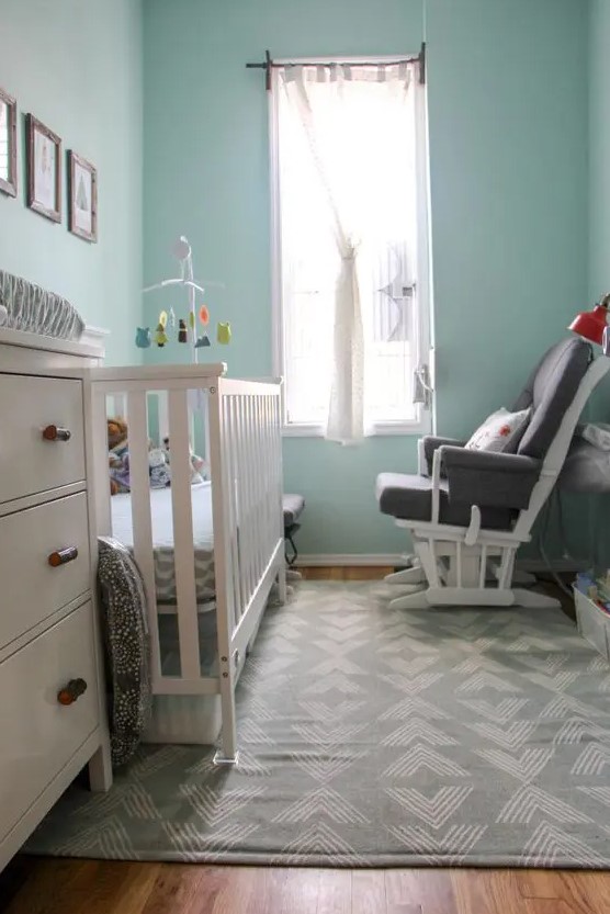 a tiny modern nursery with light blue walls, a white crib and a dresser, a grey chair, a gallery wall and printed textiles