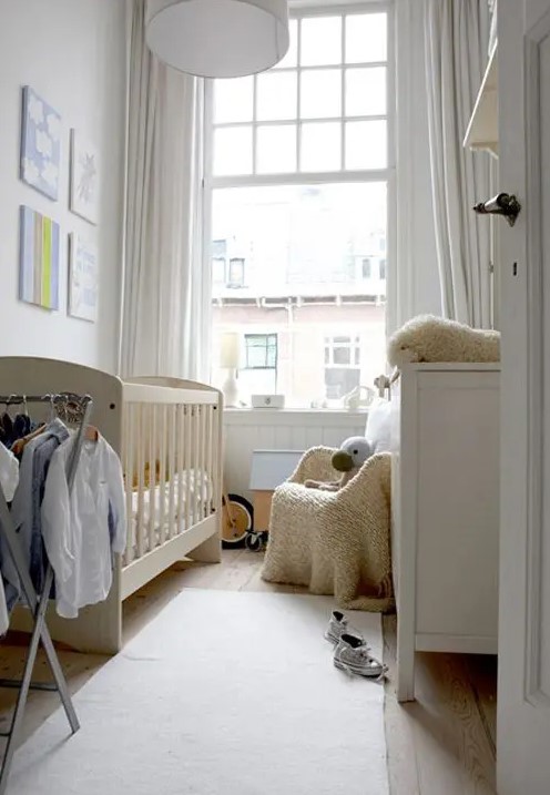 a tiny white nursery with a neutral crib, a white dresser, a neutral chair, a makeshift closet and some artwork and toys