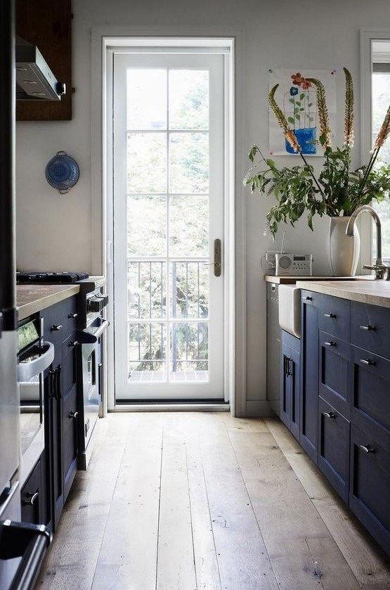 a welcoming navy farmhouse kitchen with navy cabinets and wooden countertops, with a wooden floor and much natural light from the door
