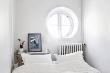 a white narrow bedroom with a porthole window, a bed with neutral bedding, a narrow nightstand at the headboard and a radiator
