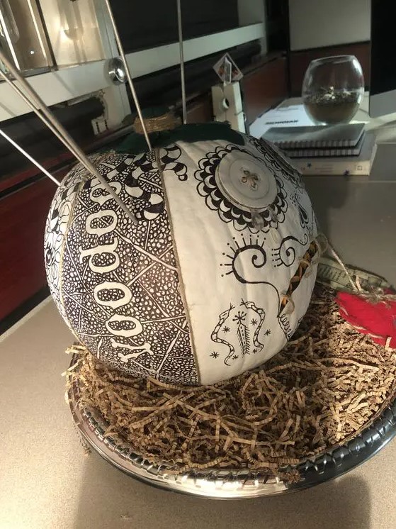 a white pumpkin decorated with a black sharpie with words, patterns and snakes and with some metal sticks is a unique solution