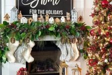 a Christmas mantel with an evergreen and snowy pinecone garland, mini houses and stockings plus a Christmas sign with matching decor