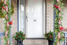a bright Christmas porch with an evergreen garland with lights, red ornaments, plaid ribbons and bows, a red rug, candle lanterns and arrangements