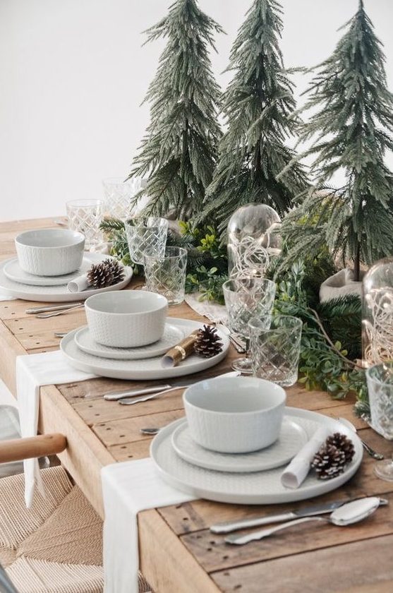 a cozy Christmas tablescape with mini trees, evergreens and greenery, white porcelain and pinecones plus white napkins