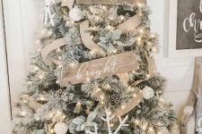 a farmhouse Christmas tree with wooden signs, a large snowflake, white ornaments, a large burlap bow and some greenery and lights