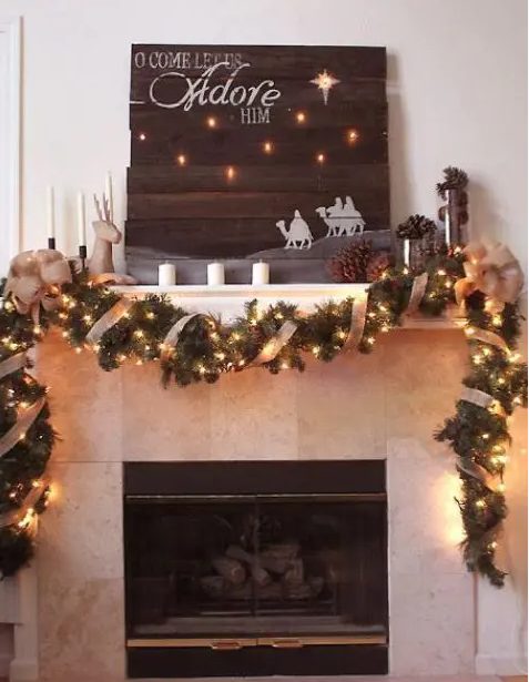 a festve mantel decorated with an evergreen garland with ribbon and lights, pinecones, candles, a large lit up sign with a religious theme