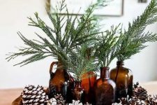 a natural woodland Christmas centerpiece of a wooden bowl with pinecones, evergreens in dark glass bottles is cool