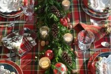 a plaid tablecloth, a greenery table runner with pinecones, apples, plaid ornaments and mercury glass candle holders