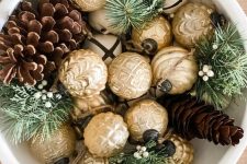a pretty Christmas centerpiece of a white bowl, gold ornaments, evergreens and pinecones is cool and easy to realize