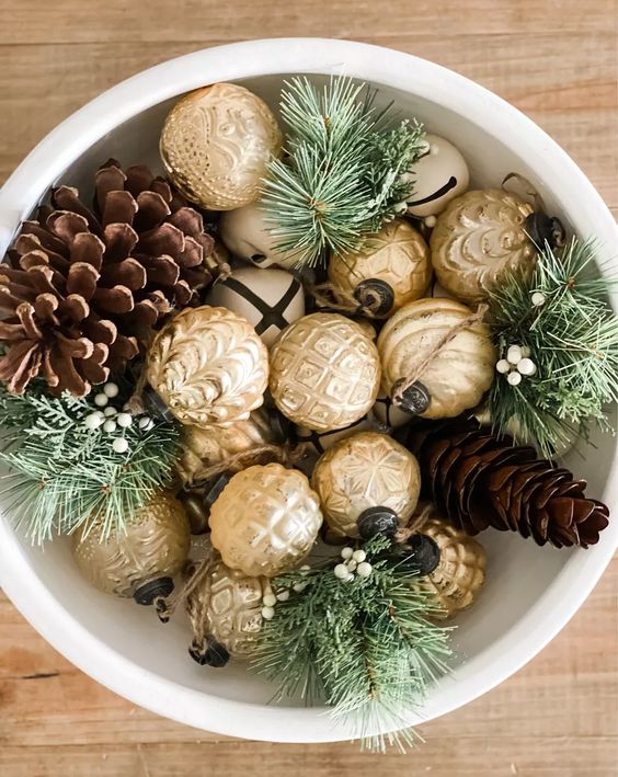 a pretty Christmas centerpiece of a white bowl, gold ornaments, evergreens and pinecones is cool and easy to realize