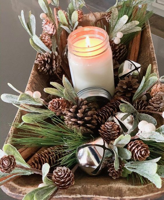 a rustic Christmas centerpiece of a wooden bowl, pinecones, greenery, bells and a candle in a jar