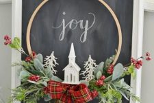 a small and cool embroidery hoop Christmas wreath with greenery, berries, a red plaid bow, a church and white faux trees