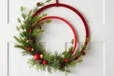 an embroidery hoop Christmas wreath in red, with evergreens, pinecones, red ornaments and wooden beads is a cool and chic idea for the holidays