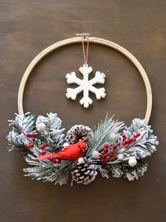an embroidery hoop Christmas wreath with snowy evergreens, berries, snowy pinecones and a red bird plus a large snowflake