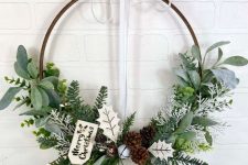 an embroidery hoop holiday wreath with evergreens, leaves, pinecones and a tag is a cool idea for the holidays