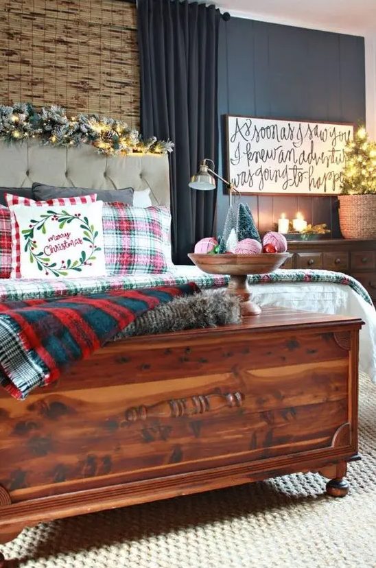 plaid bedding, a snowy evergreen and lights garland, a calligraphy sign, candles, a mini tree in a basket and a bowl with yarn balls and mini trees