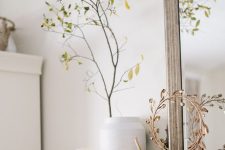 simple Scandinavian mantel decor with white pumpkins, a white vase with greenery branches and a gold wreath on a stand