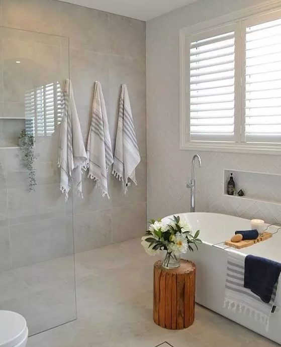 a modern neutral bathroom clad with neutral tiles, with an oval tub, a niche, printed and dark towels, some blooms