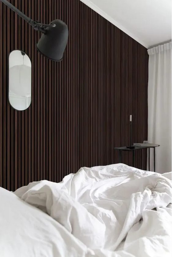 A catchy bedroom with a dark stained wood slat accent wall, a bed with neutral bedding, a black coffee table and a black sconce