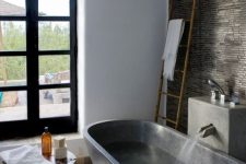 26 a moody bathroom with a white pebble tile, dark stone on the wall and a dark stone bathtub plus a wooden side table