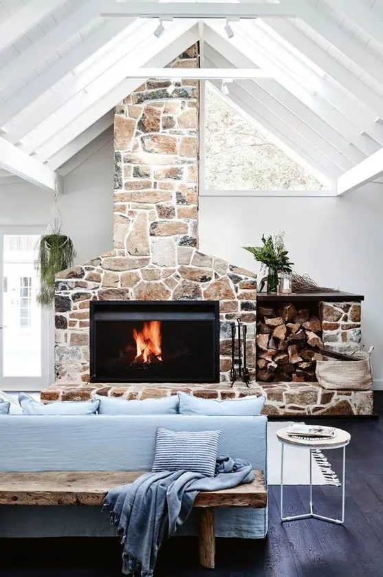 a coastal living room with a stone clad fireplace, a blue sofa, a bench, a side table and some greenery is wow