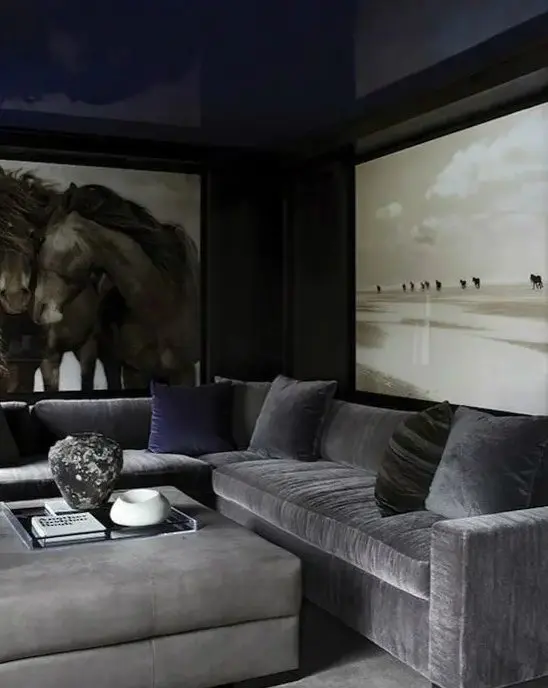 a moody living room with black walls, a navy ceiling, a grey sectional, a grey ottoman, oversized artwork and navy pillows