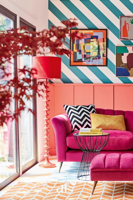 a colorful living room with a striped wall, a coral sideboard, a fuchsia sofa and ottoman, colorful artwork and a red lamp