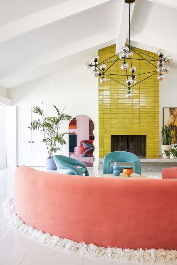 A mid century modern living room with a fireplace clad with yellow tile, aqua blue chairs, a curved coral sofa and potted plants