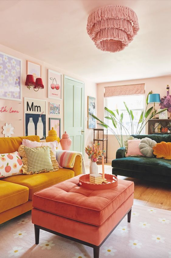 a living room with blush walls, a gallery wall, a dark green and yellow sofa, a coral ottoman and a pink tassel chandelier