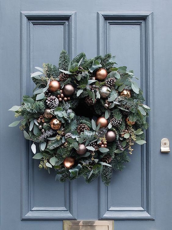 a beautiful and lush holiday wreath of evergreens and eucalyptus, copper and brown ornaments and pinecoens is chic