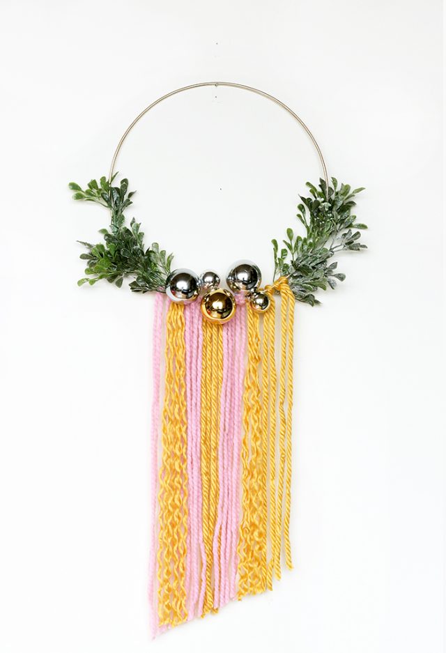 a catchy and bright boho Christmas wreath with greenery, silver ornaments and colorful yarn is a cool idea