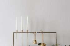 a minimal frame with a bell and an ornament, some greenery and candles in tall gold candleholders