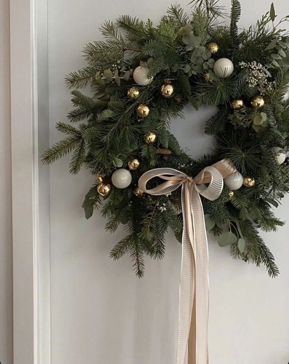 an elegant evergreen and eucalyptus Christmas wreath with white and gold ornaments and a tan ribbon bow looks cool